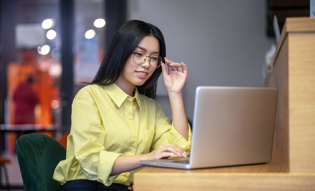 Young Asian woman with glasses working on a laptop and having coffee at her desk